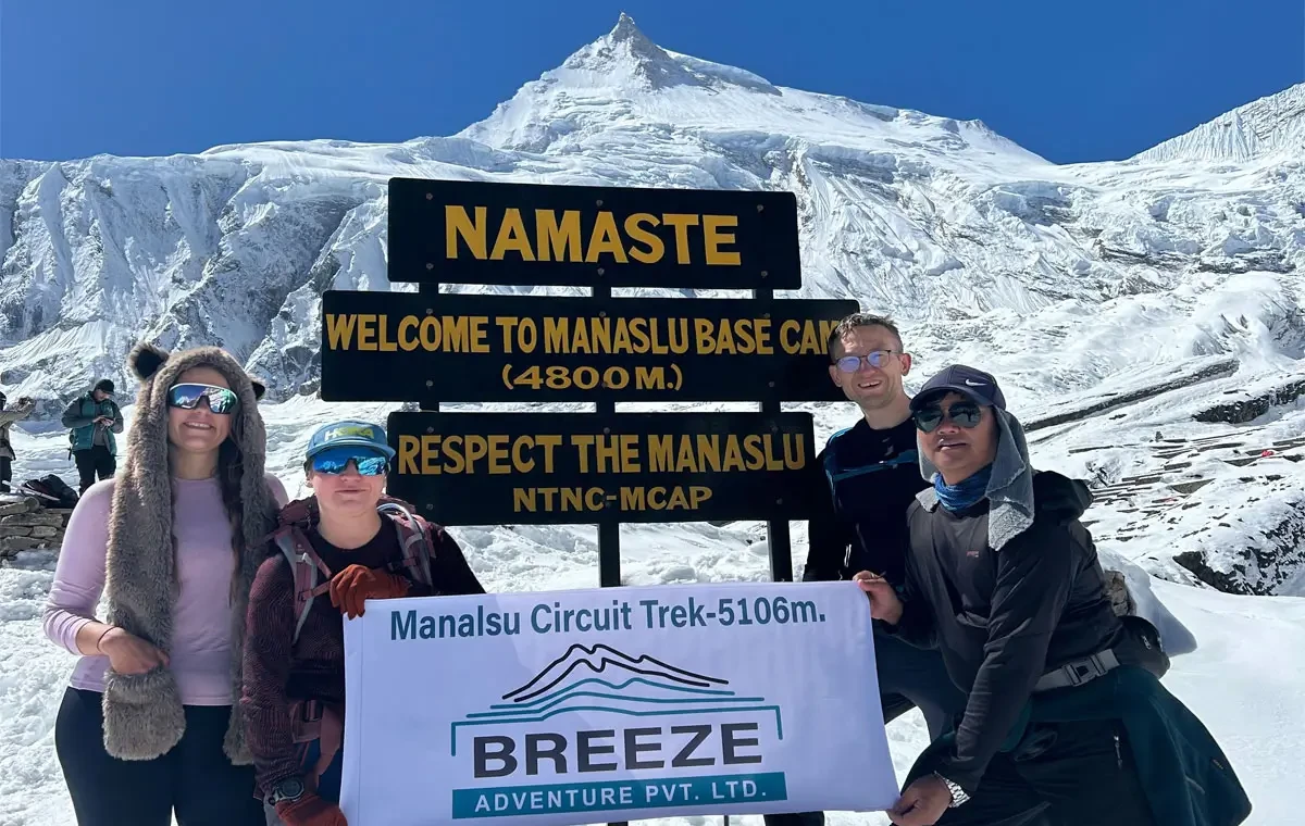 Four trekkers at the Namaste Welcome to Manaslu Base Camp 4800m signboard holding a Breeze Adventure banner below Mount Manaslu