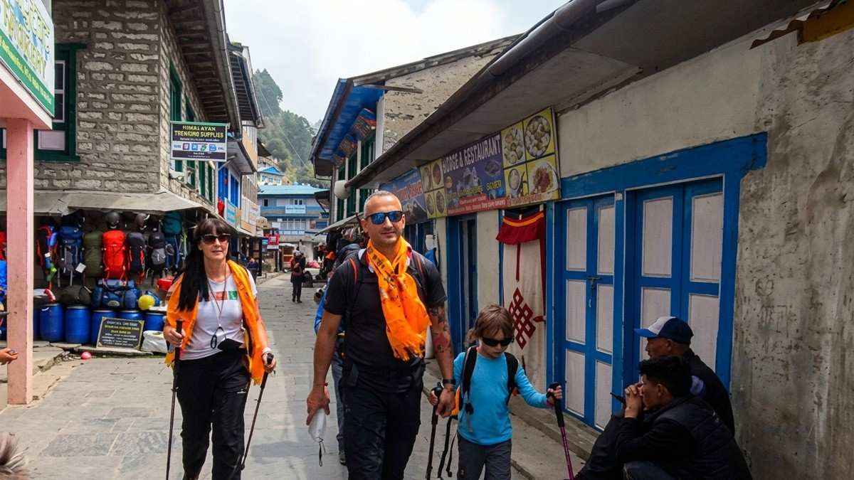 Parents and a young child with trekking poles walking past shops on the main street of Lukla, Everest regio