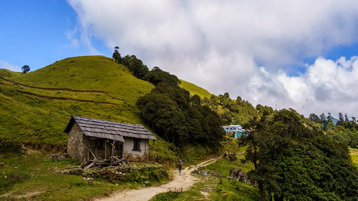 Trekker walking a trail past a traditional stone house on a green hillside in Nepal